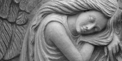 Statue of a young female angel with long hair and wings behind her as she sleeps sitting up with her head on her arm