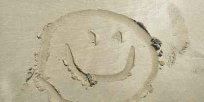 A smiley face drawn on the sand at the beach in front of a woman’s feet wearing black espadrille shoes