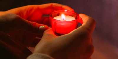 Hands holding a lit candle in a red glass candle holder in a prayerful way with another candle out of focus below.