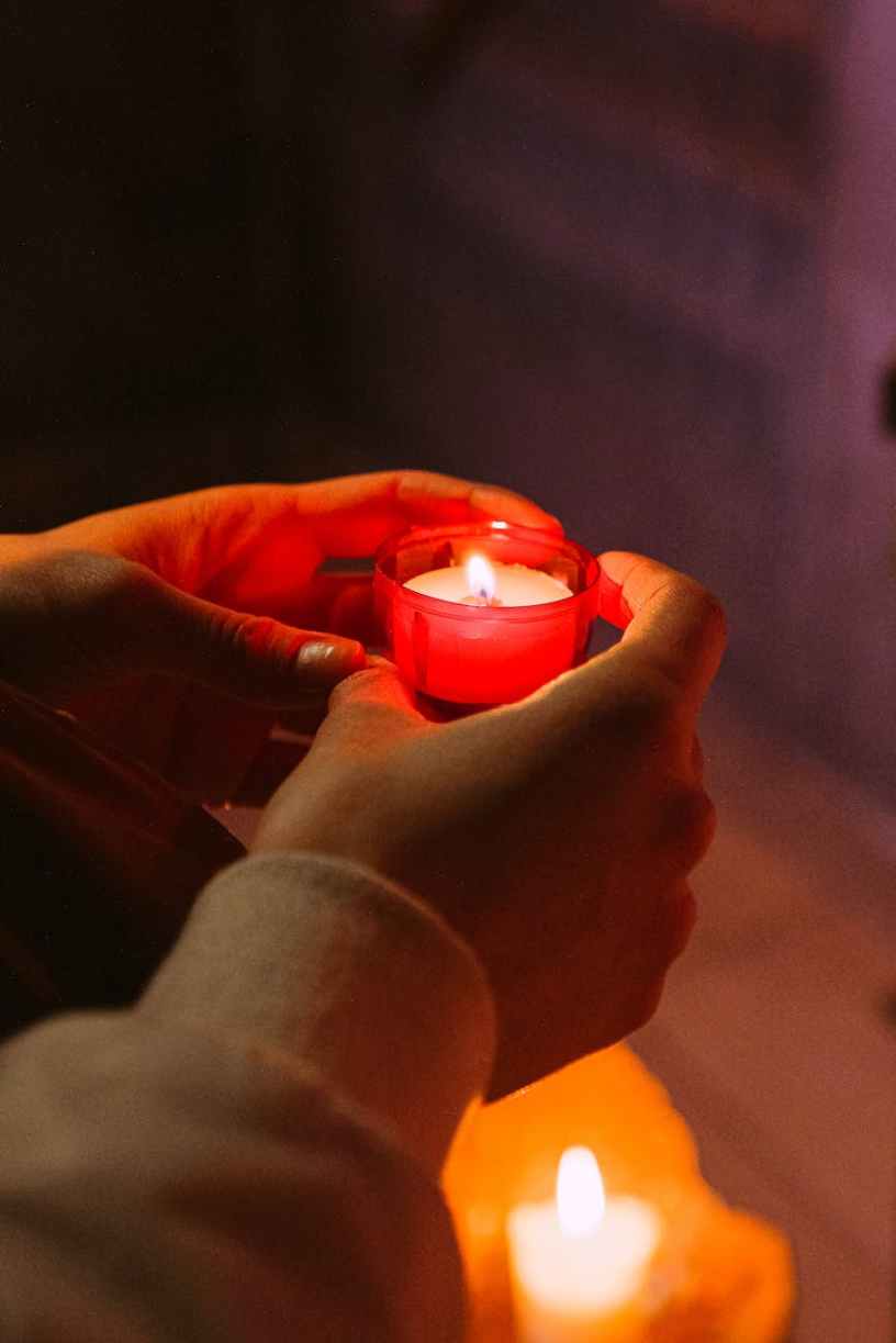 Hands holding a lit candle in a red glass candle holder in a prayerful way with another candle out of focus below.