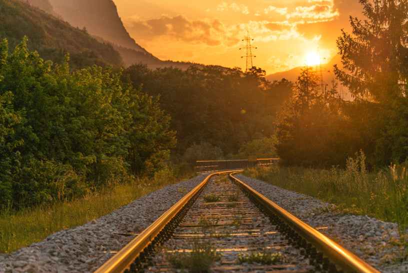 Train track with trees on either side and the sun setting in the distance.