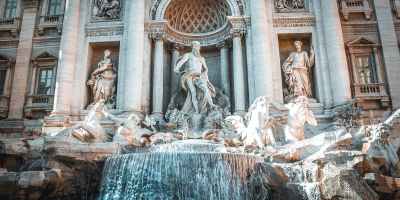 The Trevi Fountain in Rome, Italy has statues of the god Neptune with two winged horses in front of a fountain of water. In the background is a baroque style building with statues and columns.