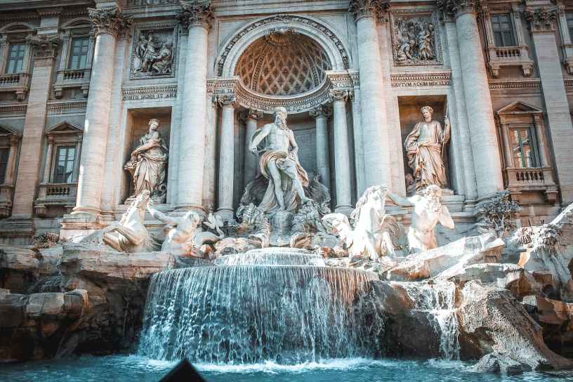 The Trevi Fountain in Rome, Italy has statues of the god Neptune with two winged horses in front of a fountain of water. In the background is a baroque style building with statues and columns.