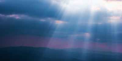 White light rays of sun shining through dark blue clouds onto a mountain range.