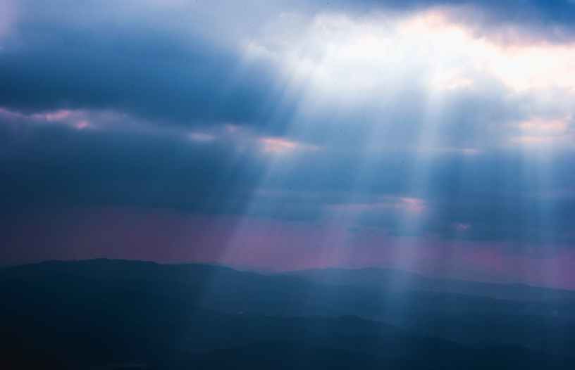 White light rays of sun shining through dark blue clouds onto a mountain range.