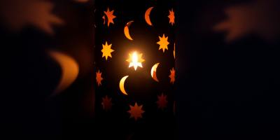 In a dark room, a lamp shade with crescent moons and stars cutout is lit and reflecting on the wall.