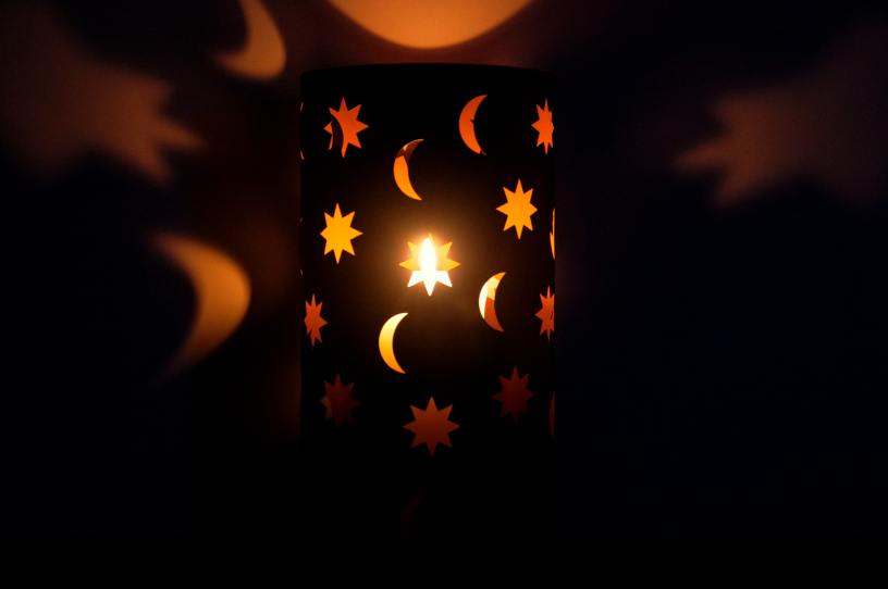 In a dark room, a lamp shade with crescent moons and stars cutout is lit and reflecting on the wall.