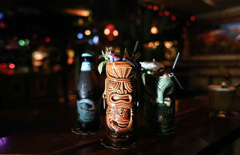 Two ceramic tiki style cocktail mugs and a beer highlighted in the foreground with blurred string lights in a bar in the background.