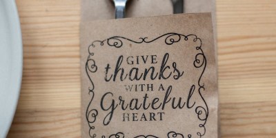 A fork and knife with a napkin sleeve that says "give thanks with a grateful heart" on a light wood table.