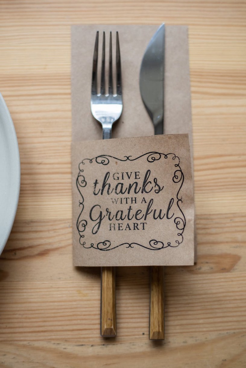 A fork and knife with a napkin sleeve that says "give thanks with a grateful heart" on a light wood table.
