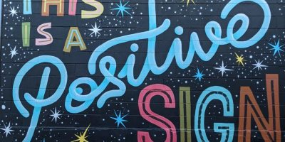 "This is a positive sign" is painted on the side of a brick building in many fun colors with star bursts.