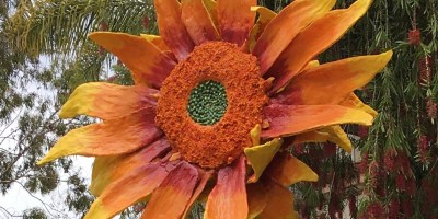 A metal sunflower sculpture with trees and roofs in the background.