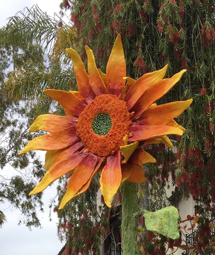 A metal sunflower sculpture with trees and roofs in the background.