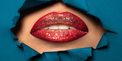 A slightly open mouth revealing white teeth with glittery red lipstick surrounded by torn teal fabric.
