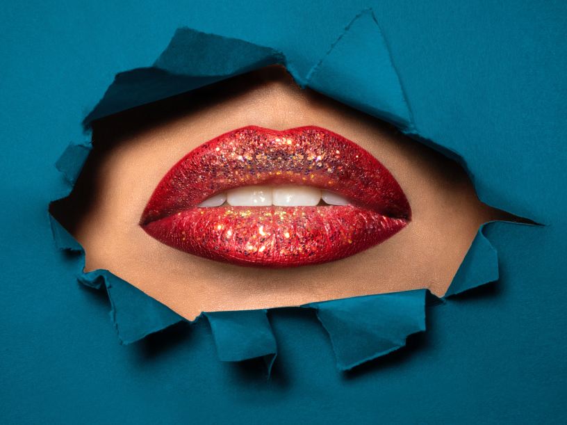 A slightly open mouth revealing white teeth with glittery red lipstick surrounded by torn teal fabric.