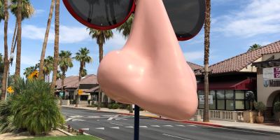 A giant nose sculpture with circular red-rimmed sunglasses outside with blue skies and palm trees in the background.