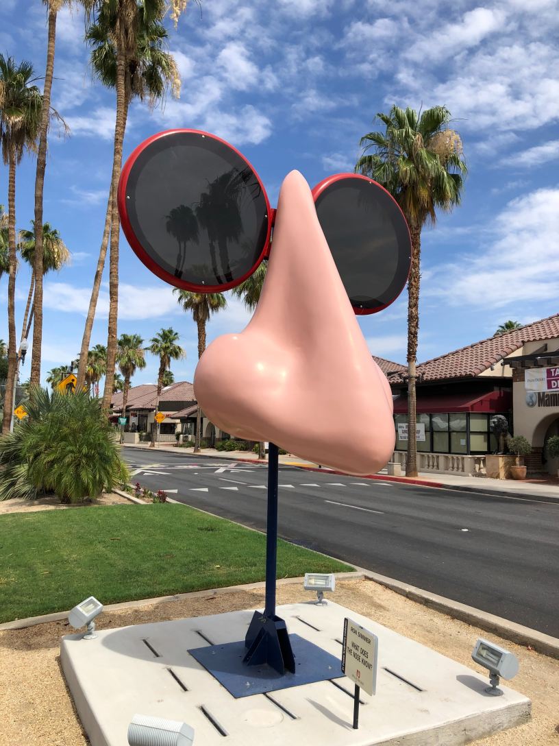 A giant nose sculpture with circular red-rimmed sunglasses outside with blue skies and palm trees in the background.