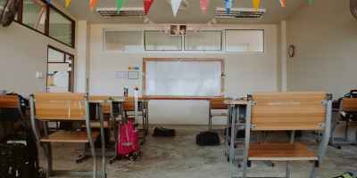 An empty classroom with two rows of desks and backpacks on the floor facing a whiteboard with colored triangle flags hanging above.