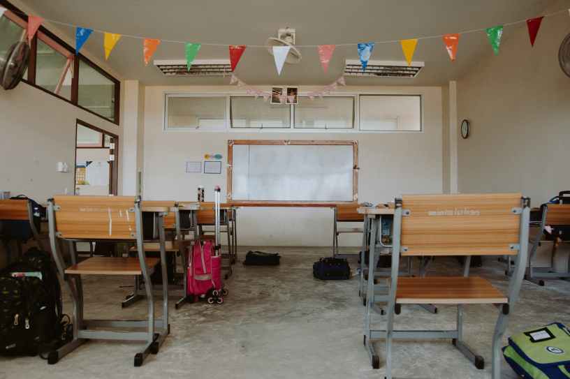 An empty classroom with two rows of desks and backpacks on the floor facing a whiteboard with colored triangle flags hanging above.