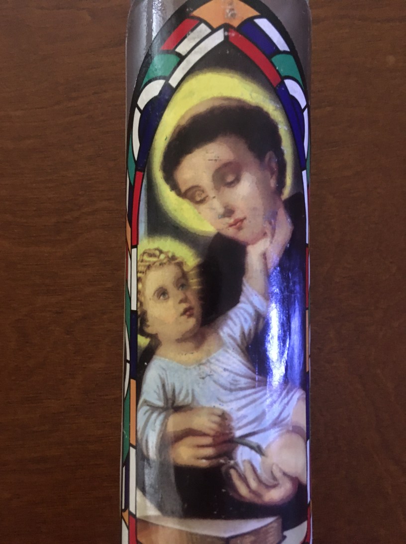 A tall glass candle with an image of baby Jesus being held by St Anthony with a woodgrain background.