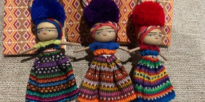 Three Guatemalan worry dolls, one with blue hair, one with purple hair and one with red hair, rest against a linen background.