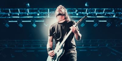 Dave Grohl of Foo Fighters plays his blue electric guitar onstage with stage lighting behind him.