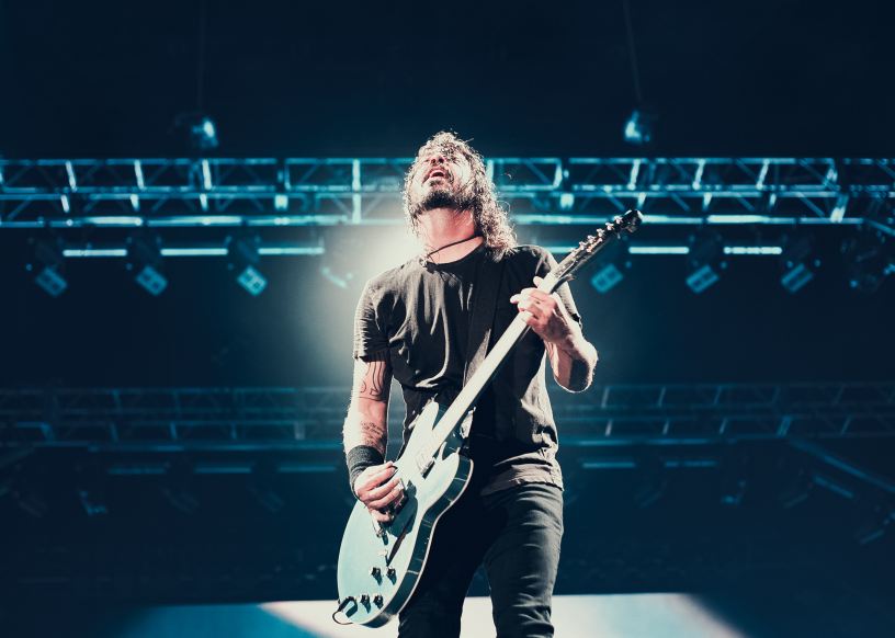 Dave Grohl of Foo Fighters plays his blue electric guitar onstage with stage lighting behind him.