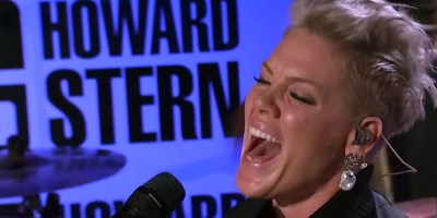 Singer and entertainer Pink sings into a microphone with a sign behind her to the left that says Howard Stern