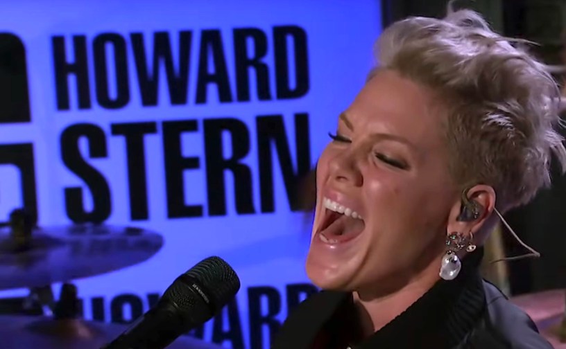 Singer and entertainer Pink sings into a microphone with a sign behind her to the left that says Howard Stern