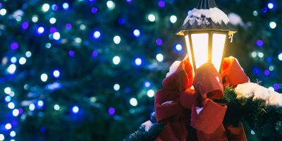 A decorative lighted lamp post with a big red bow is dusted with snow in front of blurred purple, blue and white lights in a green tree.