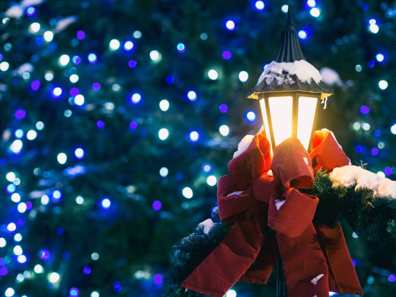 A decorative lighted lamp post with a big red bow is dusted with snow in front of blurred purple, blue and white lights in a green tree.
