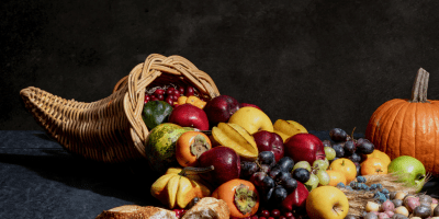 A cornucopia with colorful grapes, apples, persimmons, carambola and potatoes spilling out has a broken loaf of bread in front.