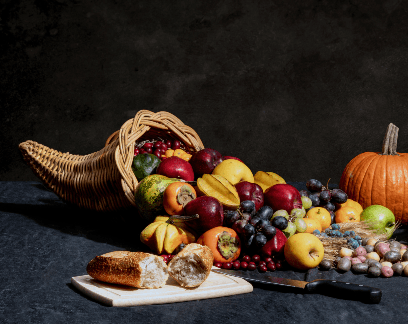 A cornucopia with colorful grapes, apples, persimmons, carambola and potatoes spilling out has a broken loaf of bread in front.