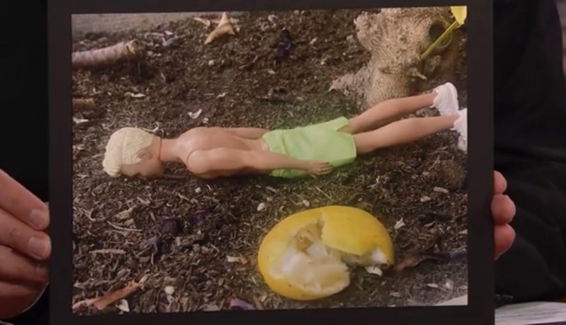 A photograph of a blonde Ken doll face down on the ground beside a squashed lemon.