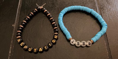 Two friendship bracelets from a Taylor Swift Eras Tour concert. The friendship bracelet on the left is black and gold beads with REPUTATION and the friendship bracelet on the right has aqua rings with 1989.