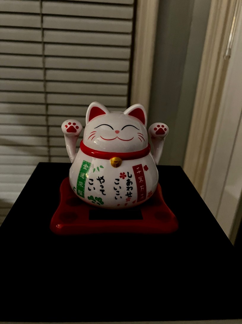 Japanese lucky cat, called Maneki Neko, is a rotund white cat with red accents and both paws raised in the air.