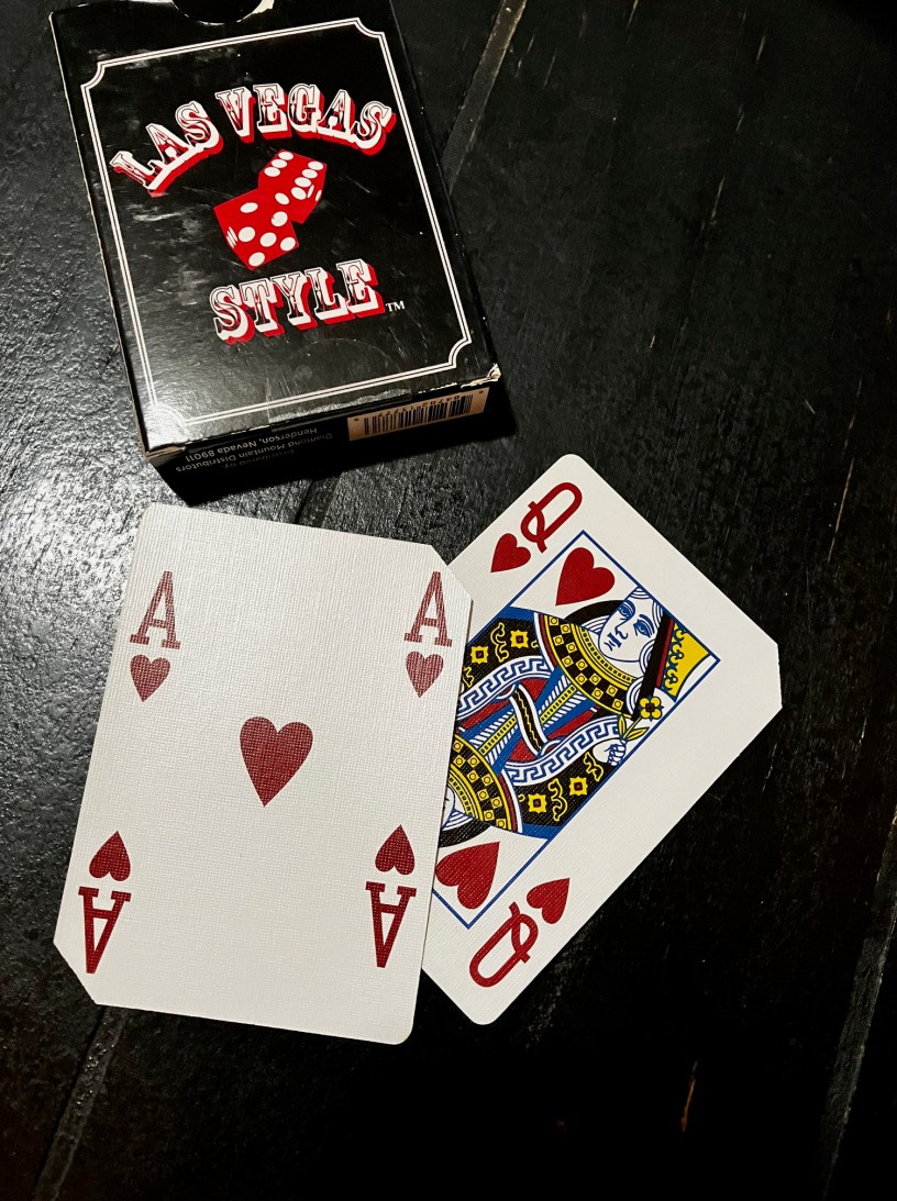 A black box that says Las Vegas Style is above two playing cards, a red ace of hearts and a red queen of hearts on a black surface.