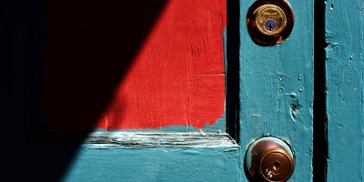 An aqua painted old door with red swatch in the shadow has a metal deadbolt lock above a door knob.
