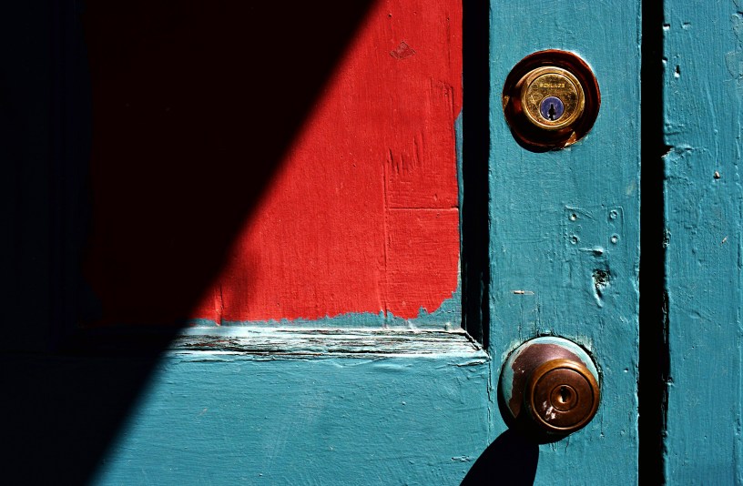 An aqua painted old door with red swatch in the shadow has a metal deadbolt lock above a door knob.