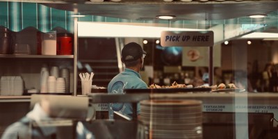 A pick-up area at a restaurant has paper towels across a top shelf above a man in a blue shirt looking away from plates of food on a counter under a sign that says "pick up here."