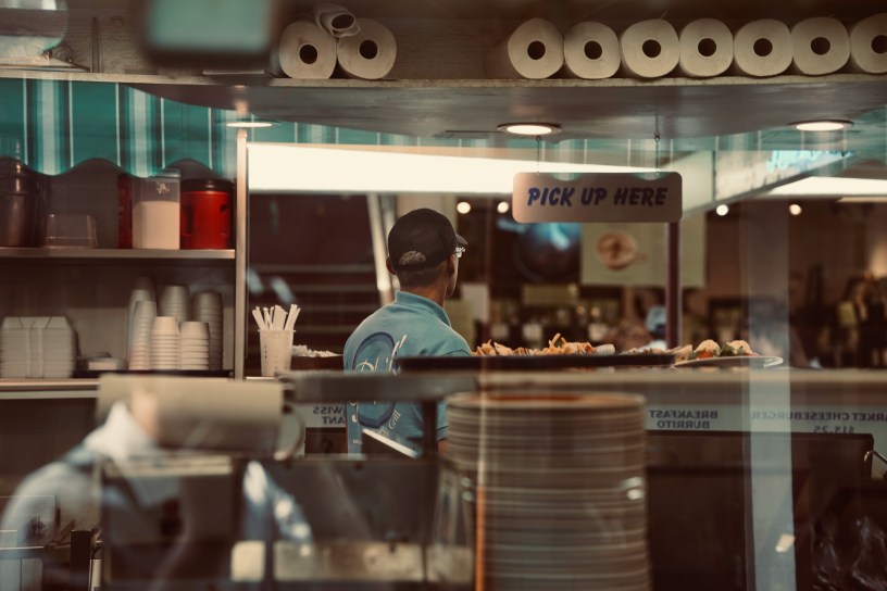 A pick-up area at a restaurant has paper towels across a top shelf above a man in a blue shirt looking away from plates of food on a counter under a sign that says "pick up here."