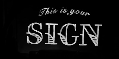 A black chalkboard with the words "This is your" written in script on the top line and the word "sign" on the bottom line written in decorative all caps with white chalk.