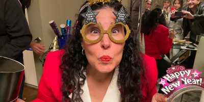 Woman with dark wavy hair wearing a red jacket with white collar and new year's eve glasses and tiara with party-goers reflected in a mirror behind her.