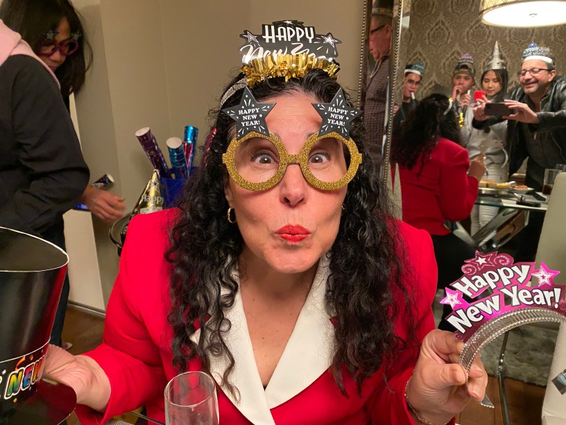 Woman with dark wavy hair wearing a red jacket with white collar and new year's eve glasses and tiara with party-goers reflected in a mirror behind her.